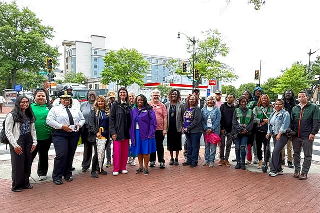 Photo of CM Brooke Pinto (in pink pants) and CM Janeese Lewis George (in purple jacket) in front of a semi-circle of community members, MPD, Safe Passage Team members.